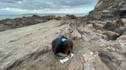 Image of Ruth Broadbent drawing on rocky beach with sea in background (2021)