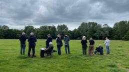 A row of people (sitting and standing) sketching in a park. Grassy area looking towards trees.